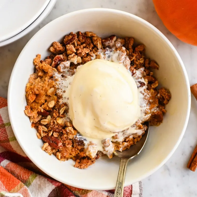 Pumpkin crisp served warm in a white bowl with vanilla ice cream, oat pecan topping, cinnamon sticks, a pumpkin, and a plaid towel nearby.