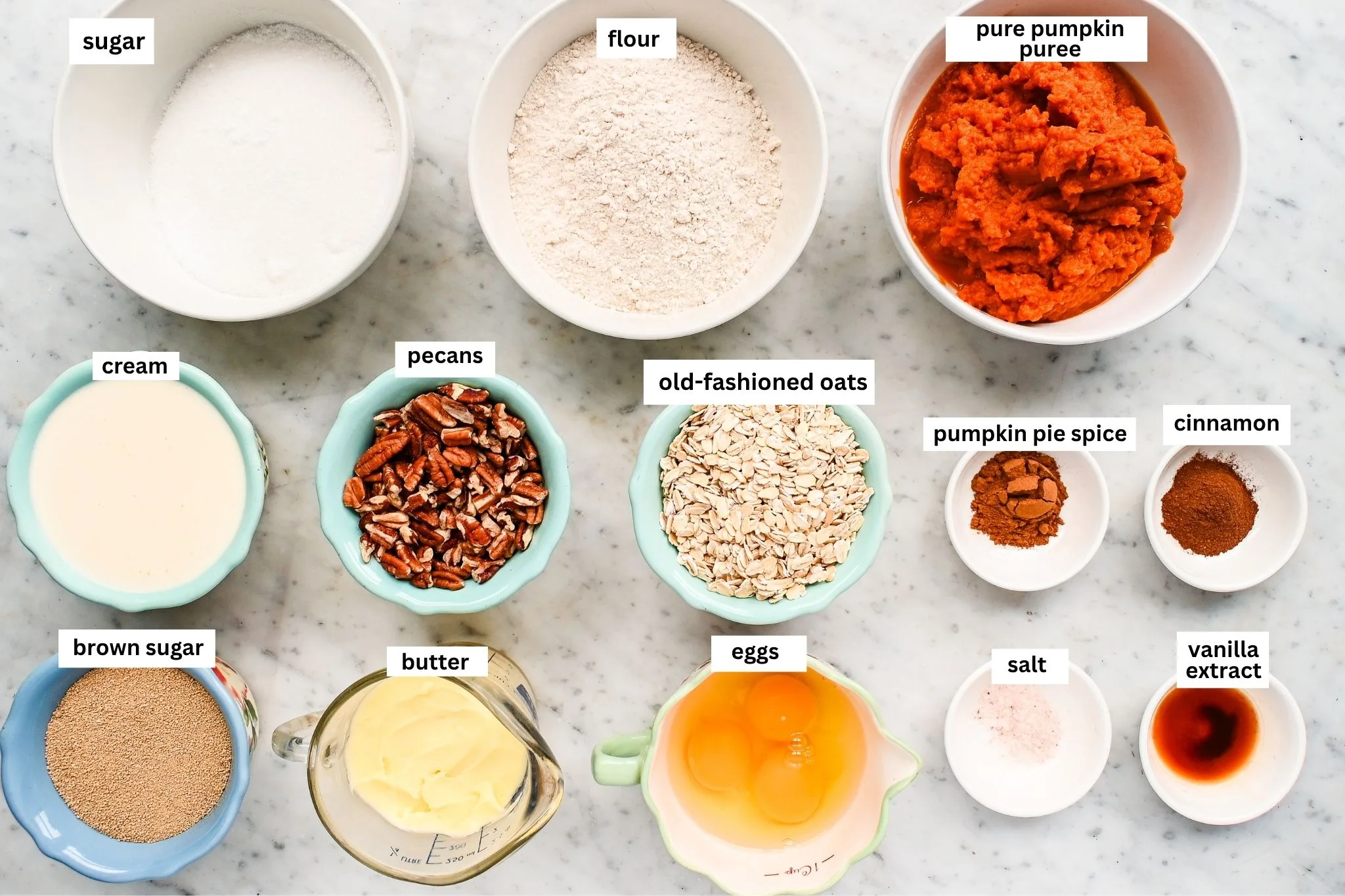 Pumpkin crisp ingredients arranged on a marble counter, including pumpkin puree, oats, pecans, flour, sugar, eggs, cream, butter, spices, vanilla, and salt.