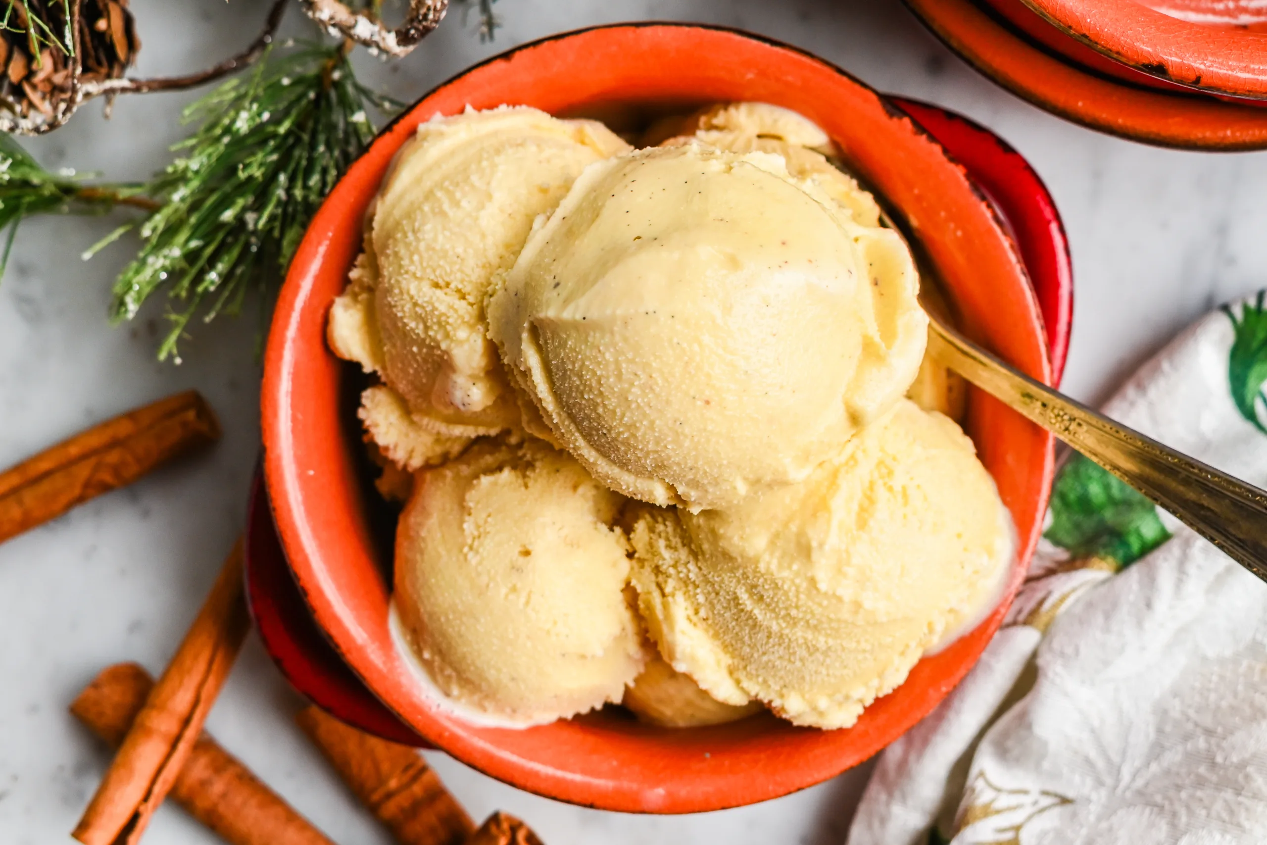 Homemade eggnog ice cream in a red bowl with cinnamon sticks, greenery, and holiday decorations nearby.