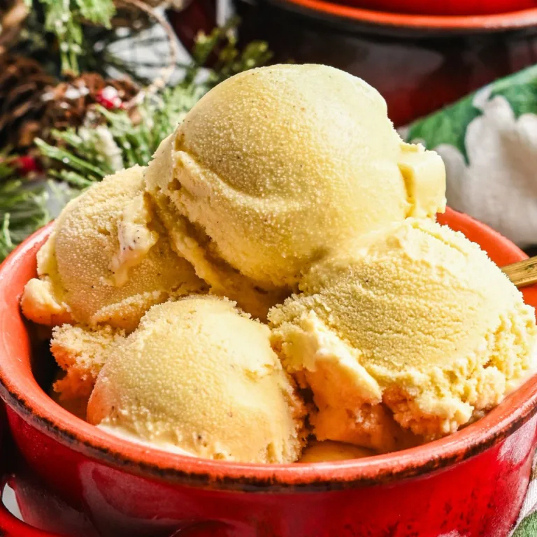 Homemade eggnog ice cream served in a red bowl with a spoon, cinnamon sticks, greenery, and a Christmas towel.