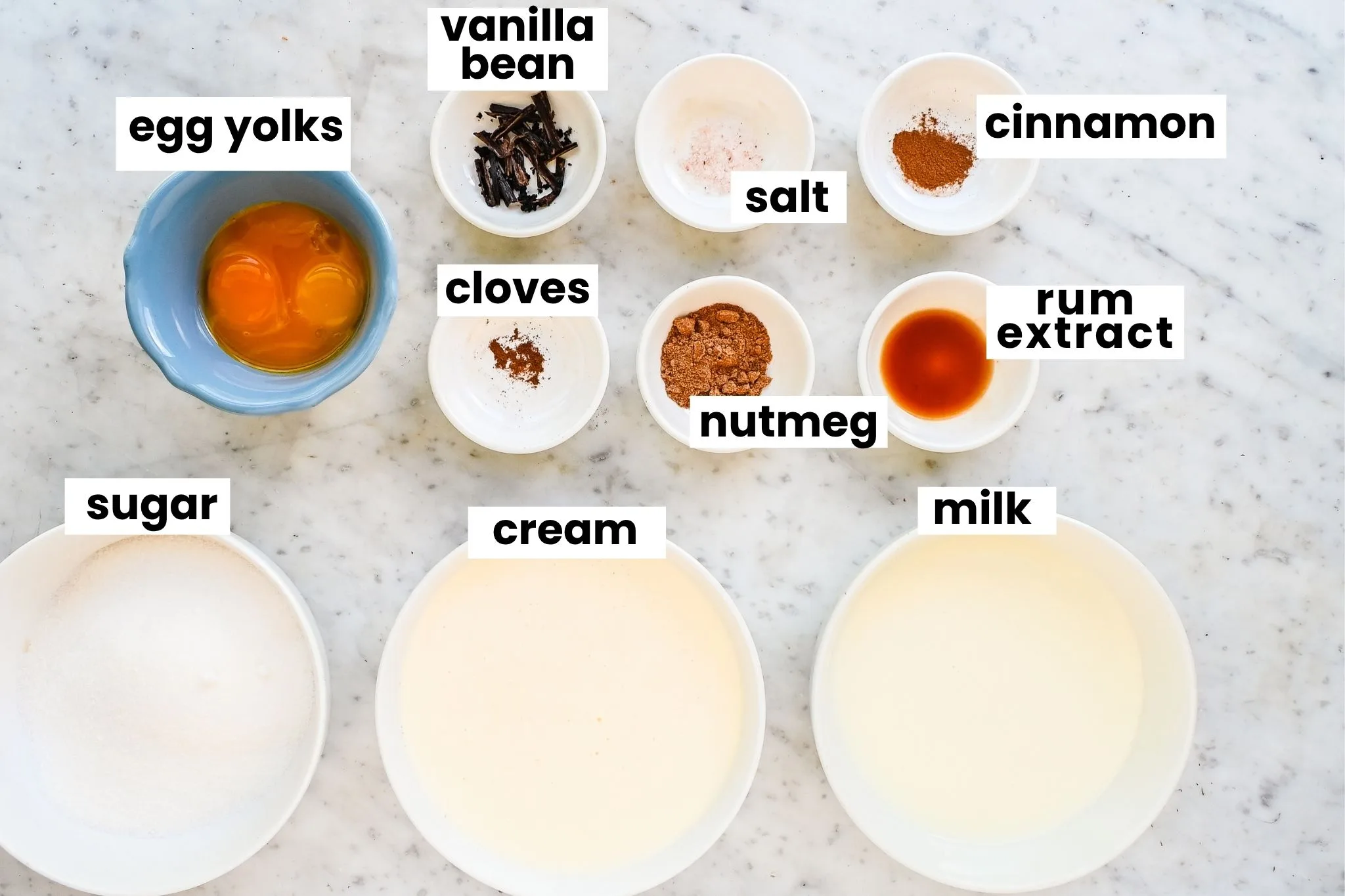 Homemade eggnog ice cream ingredients arranged on a marble counter, including egg yolks, sugar, cream, milk, vanilla, spices, and salt.