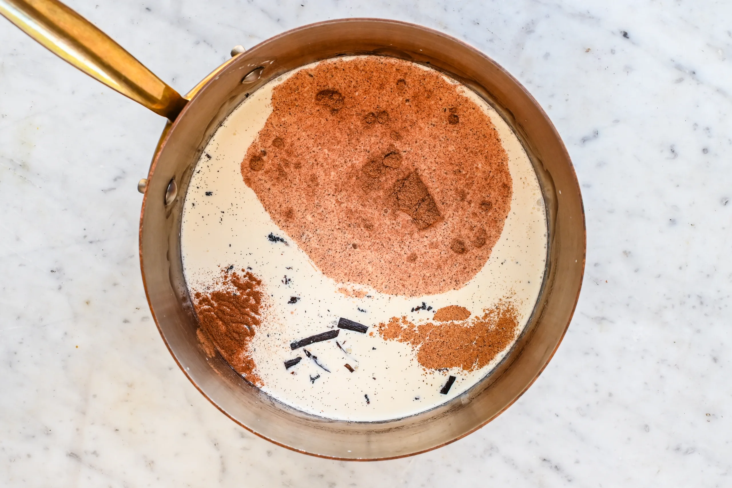 Heavy cream, milk, vanilla bean, nutmeg, cinnamon, cloves, and salt in a copper saucepan for homemade eggnog ice cream.