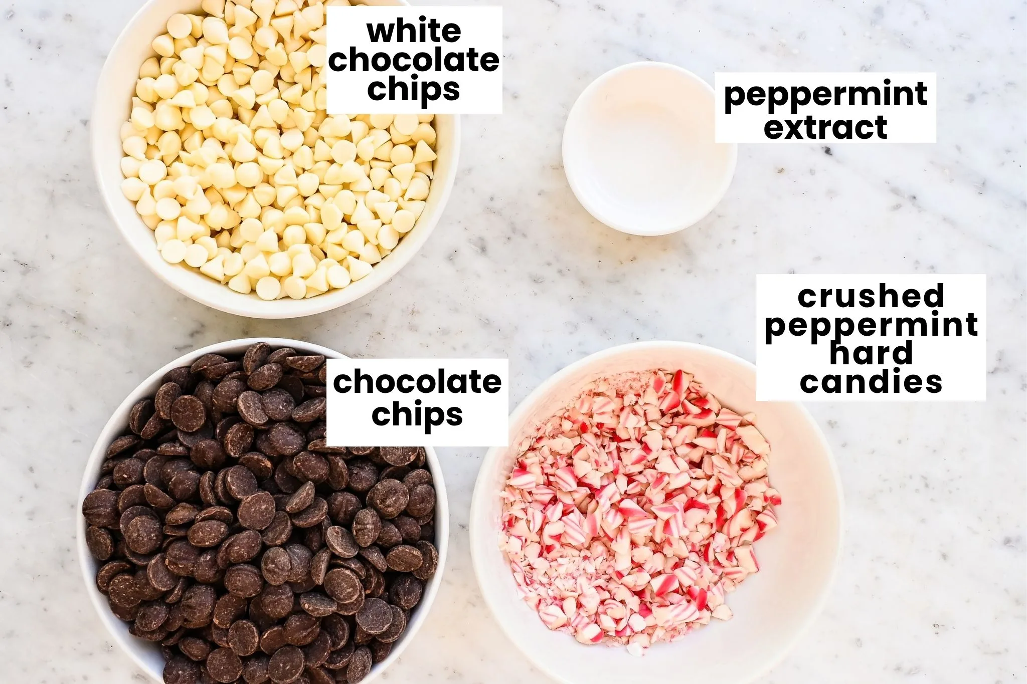 Peppermint bark ingredients arranged on a marble counter, including white chocolate chips, dark chocolate chips, crushed peppermint candy, and peppermint extract.