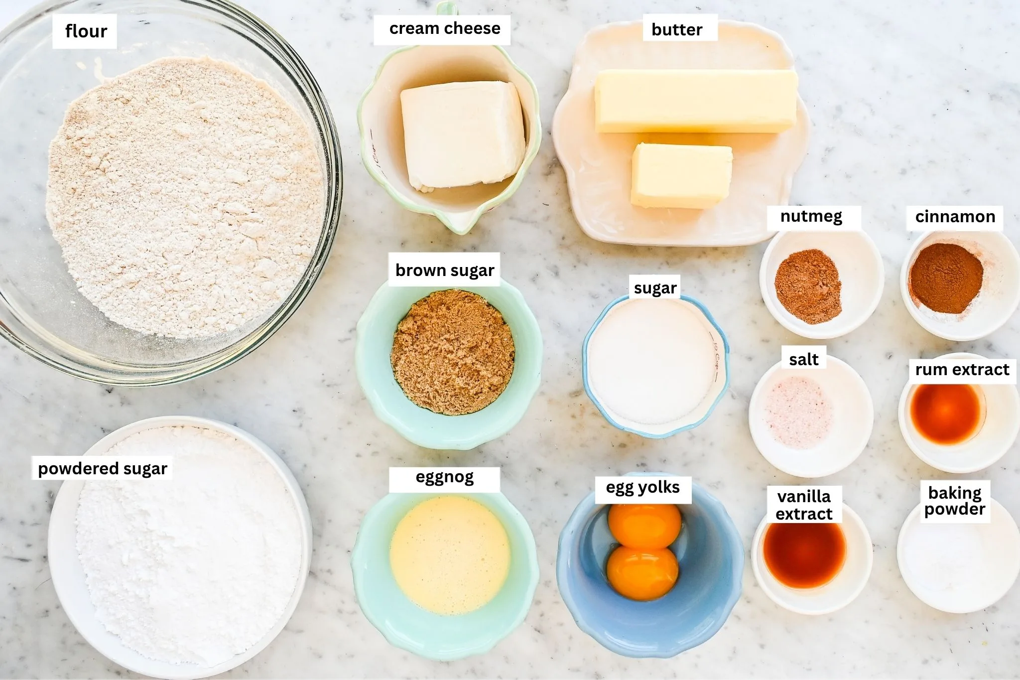 Soft eggnog cookies ingredients arranged on a marble counter, including flour, cream cheese, butter, sugars, egg yolks, eggnog, vanilla, rum extract, nutmeg, cinnamon, salt, and powdered sugar.
