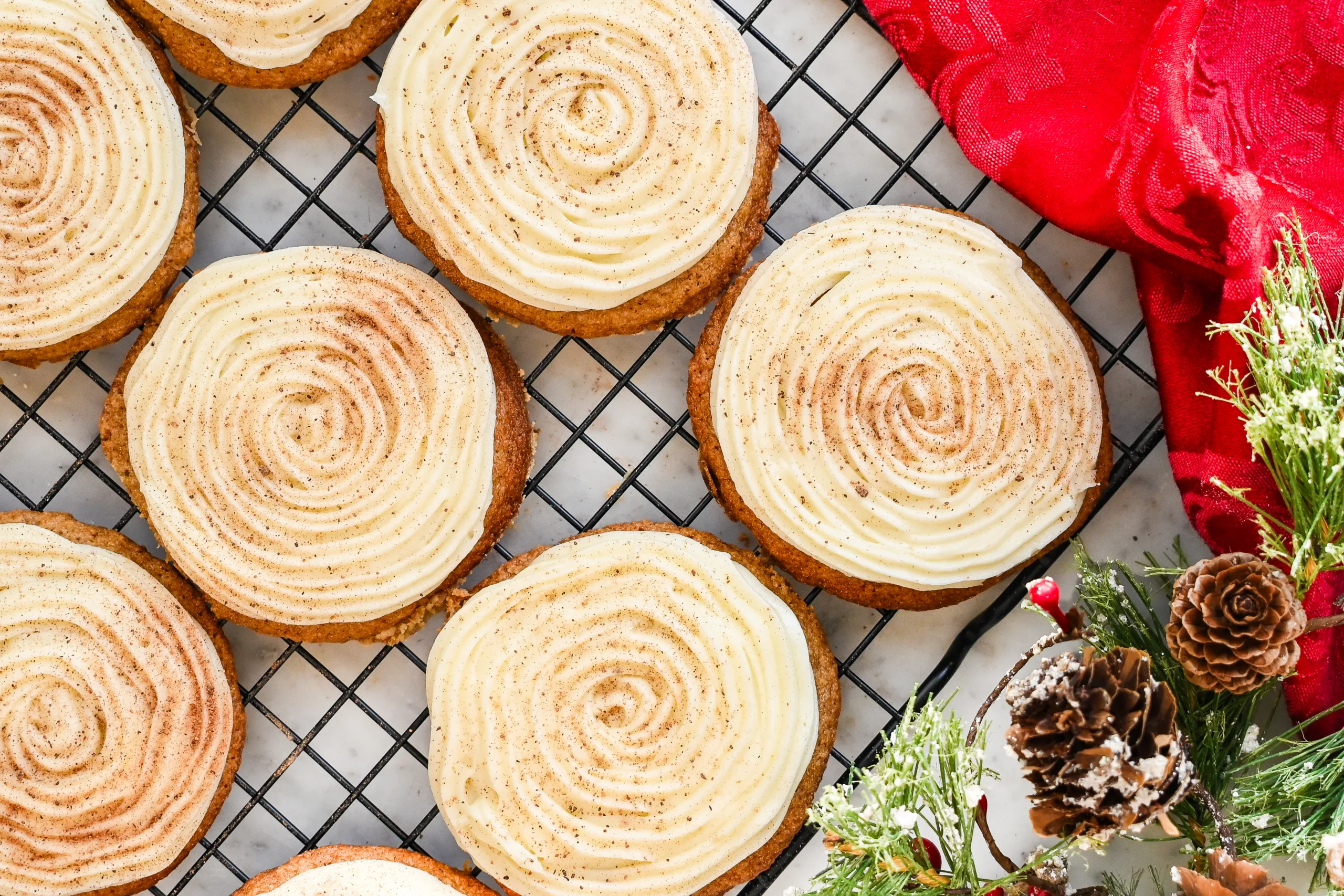 Frosted eggnog cookies on a wire rack with nutmeg, red cloth, pinecones, and greenery.