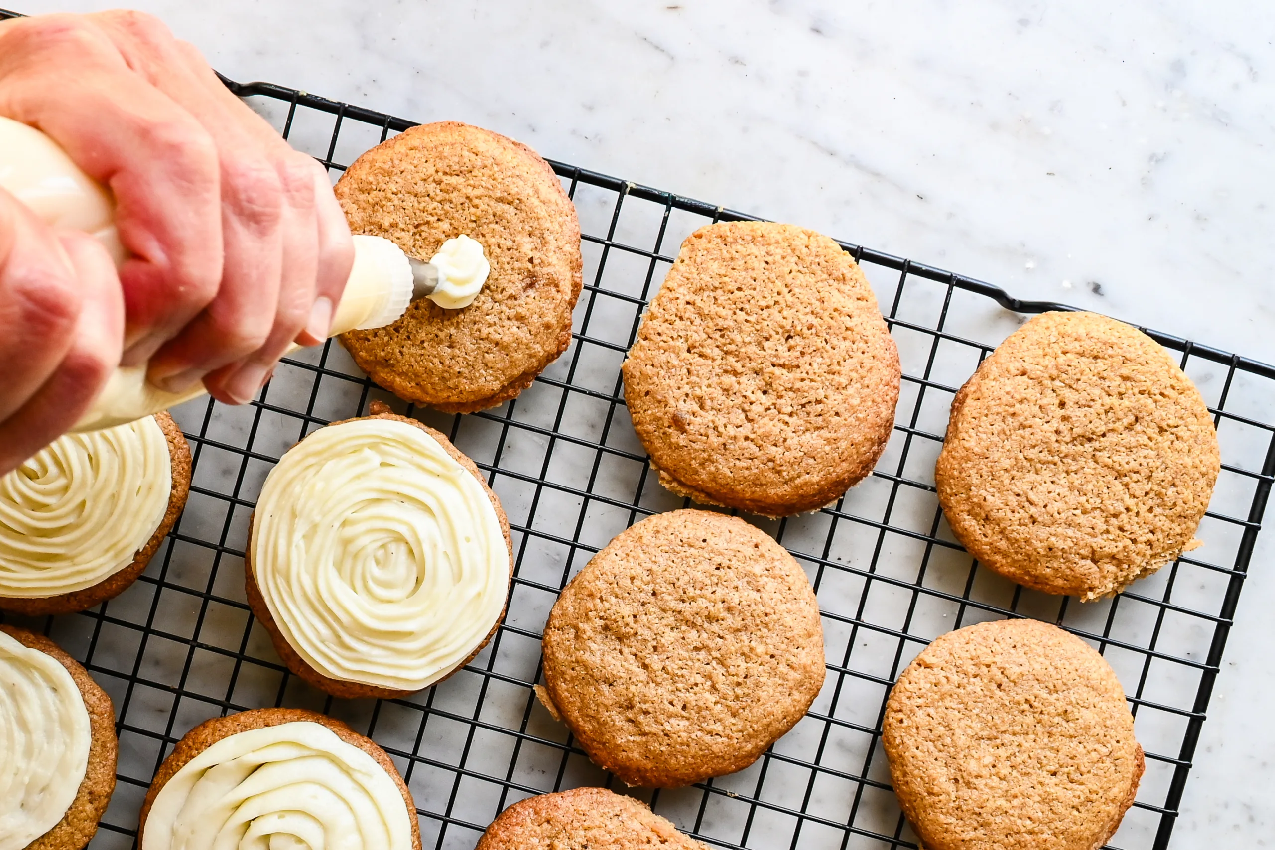 Eggnog cream cheese frosting being piped onto cooled cookies on a wire rack.