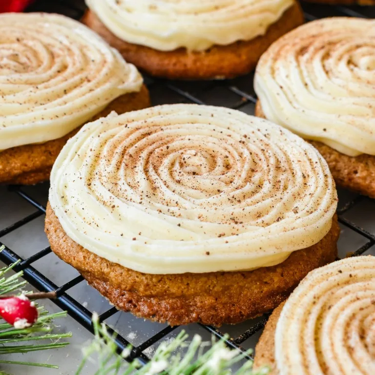 Soft eggnog cookies topped with cream cheese frosting and nutmeg on a wire rack with red cloth, pinecones, and greenery nearby.