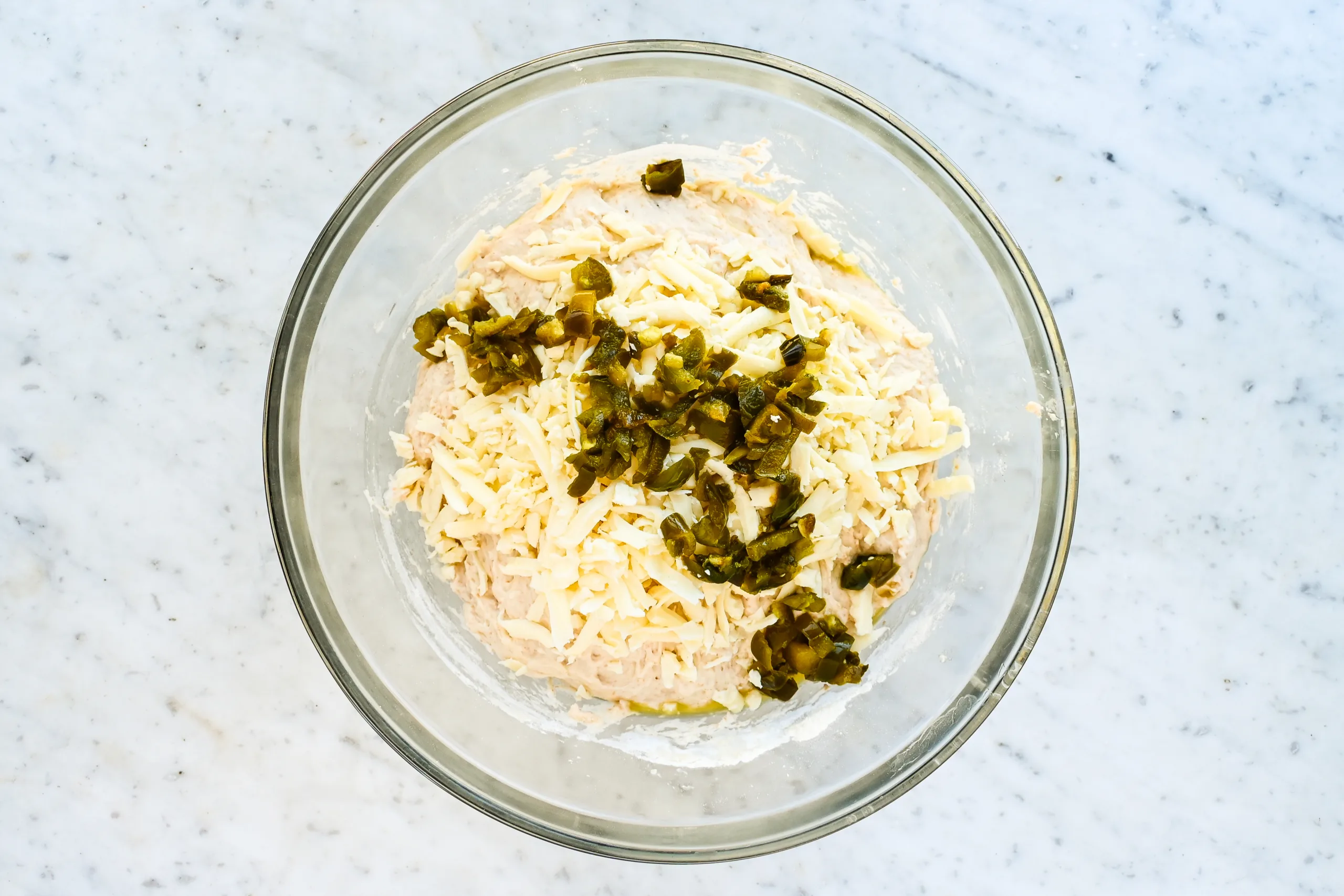 Shredded cheddar cheese and diced jalapeños added to sourdough focaccia dough in a glass mixing bowl.