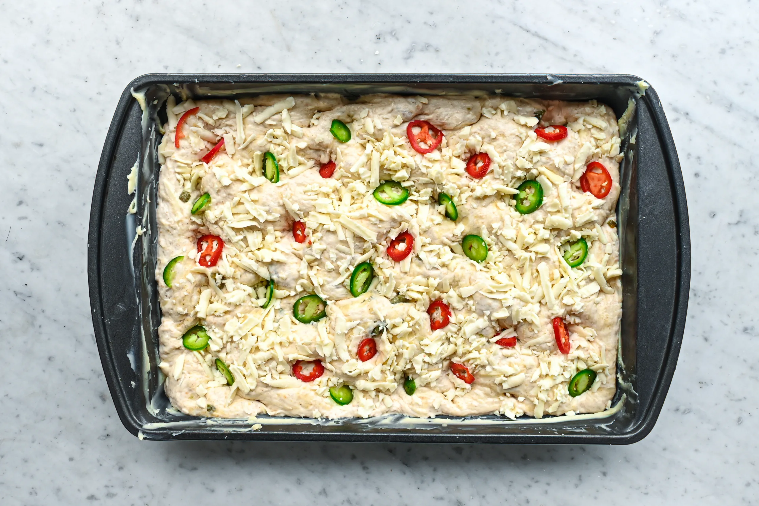 Unbaked jalapeño cheddar sourdough focaccia topped with shredded cheddar cheese and sliced red and green jalapeños.