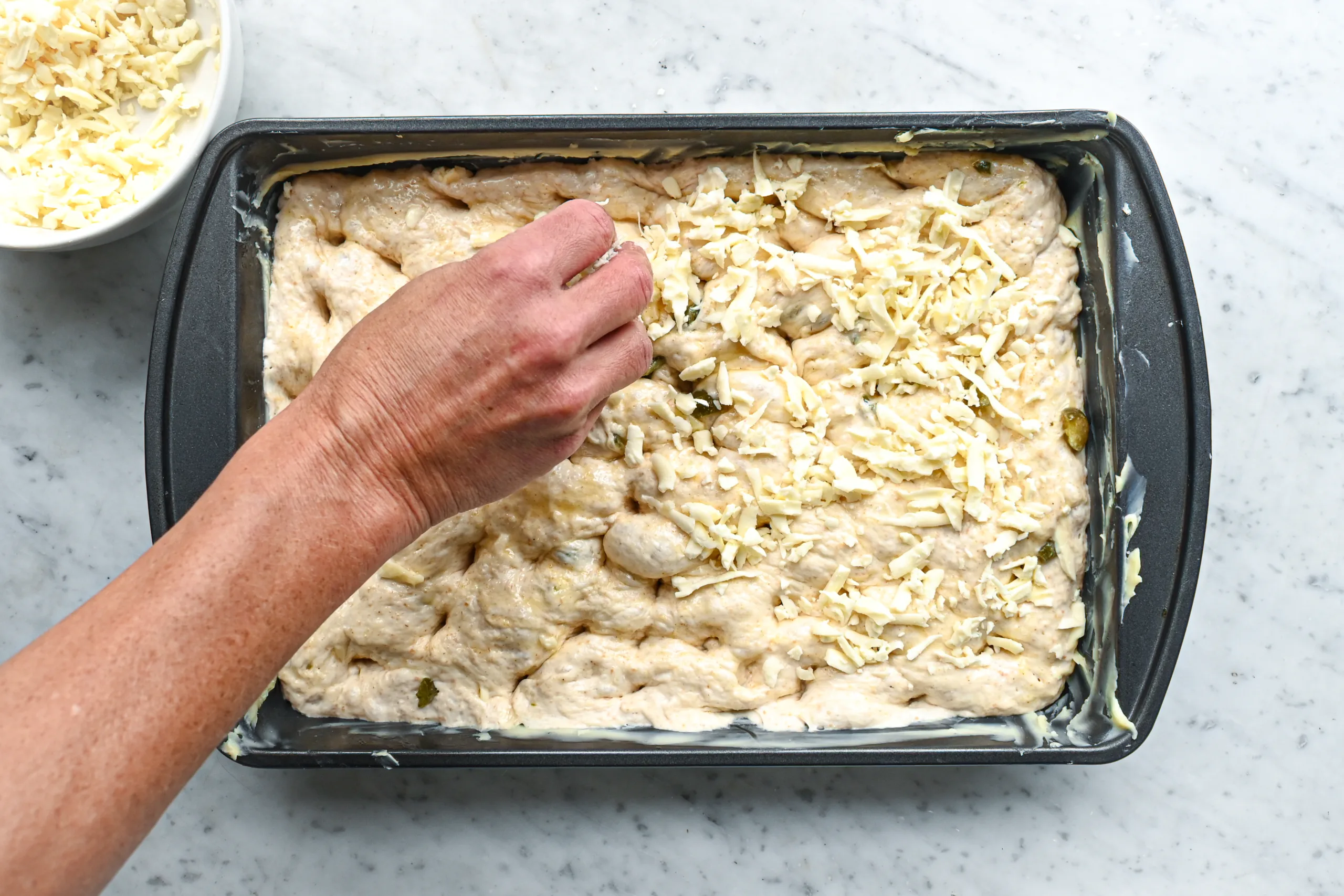 Shredded cheddar cheese being sprinkled over jalapeño cheddar sourdough focaccia dough.