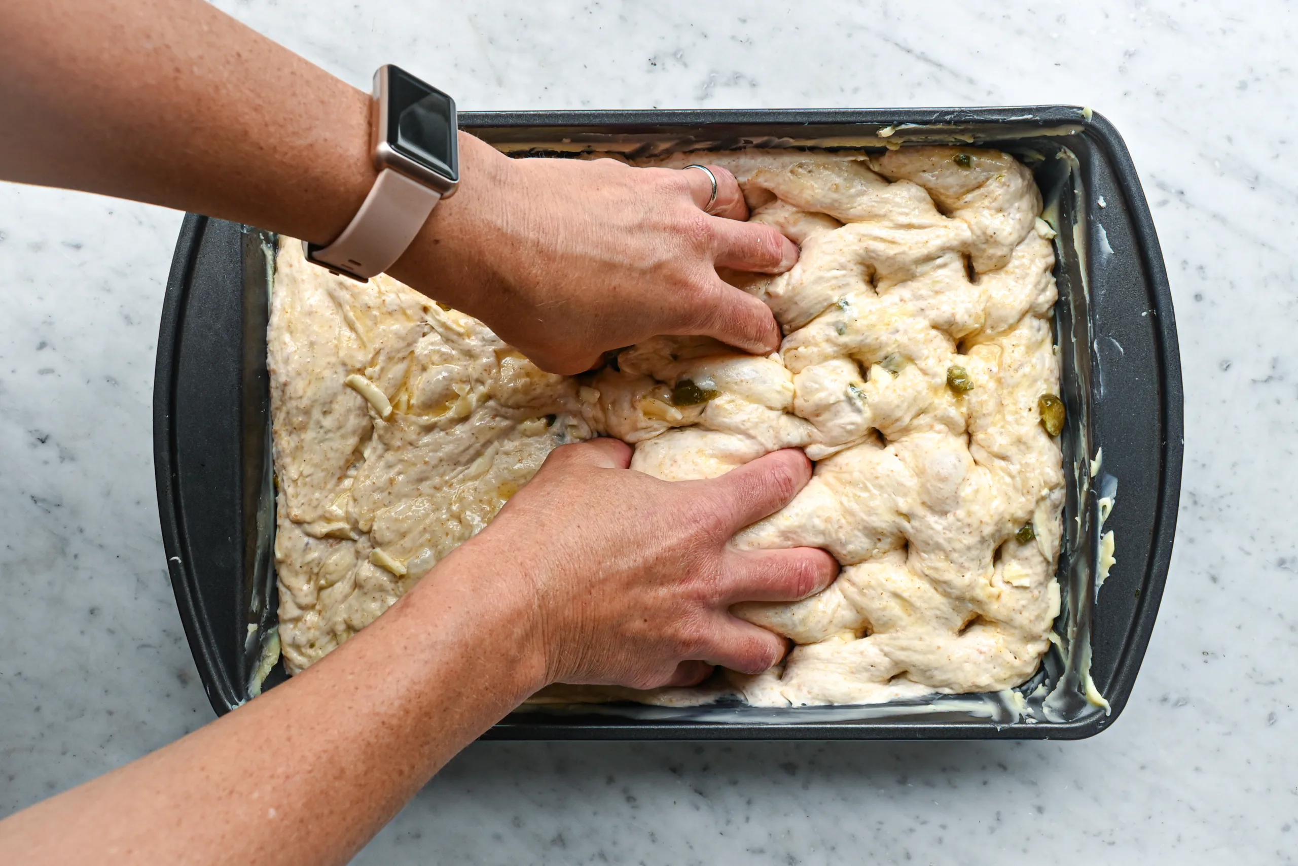 Hands dimpling jalapeño cheddar sourdough focaccia dough in a buttered baking pan.