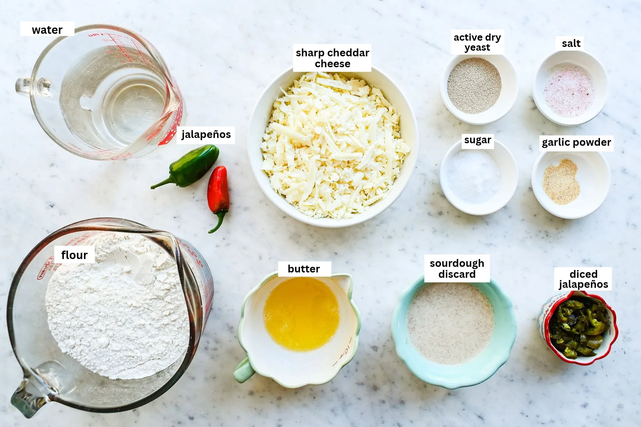 Ingredients for jalapeño cheddar sourdough focaccia arranged on a marble counter, including water, flour, sourdough discard, butter, sharp cheddar, jalapeños, yeast, sugar, salt, and garlic powder.