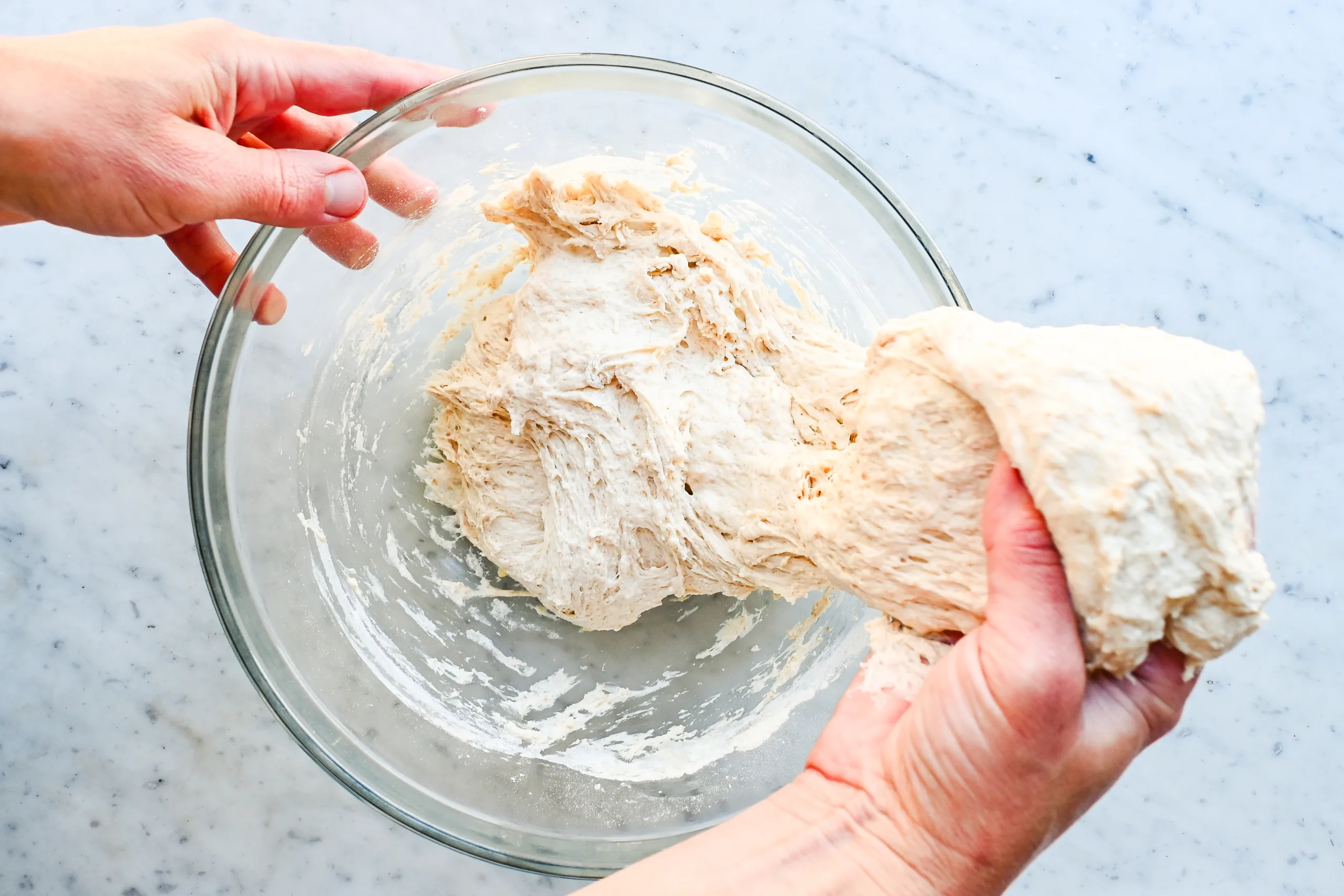 Hands stretching sticky sourdough focaccia dough in a glass mixing bowl.