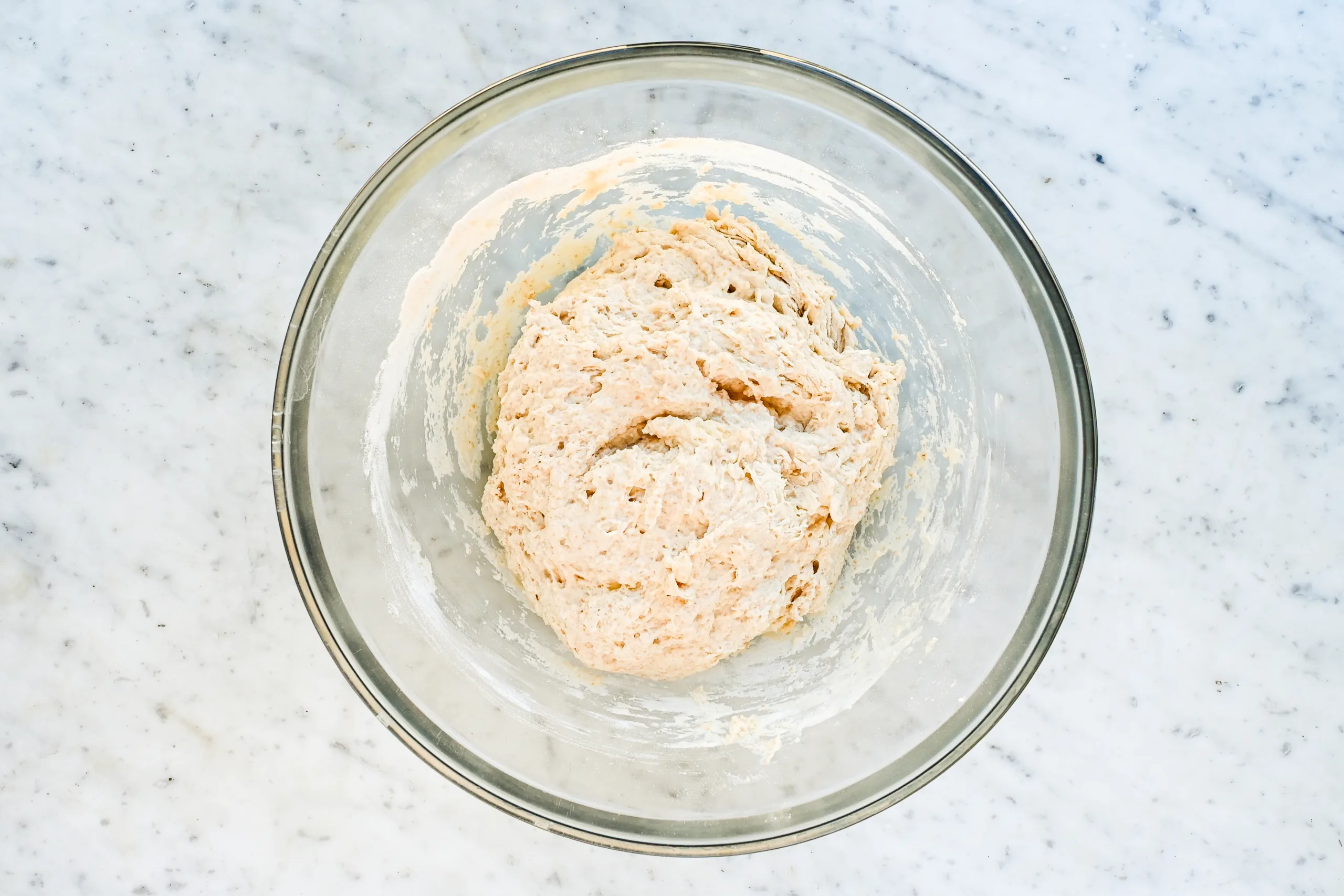 Sticky sourdough focaccia dough mixed in a glass bowl on a marble counter.