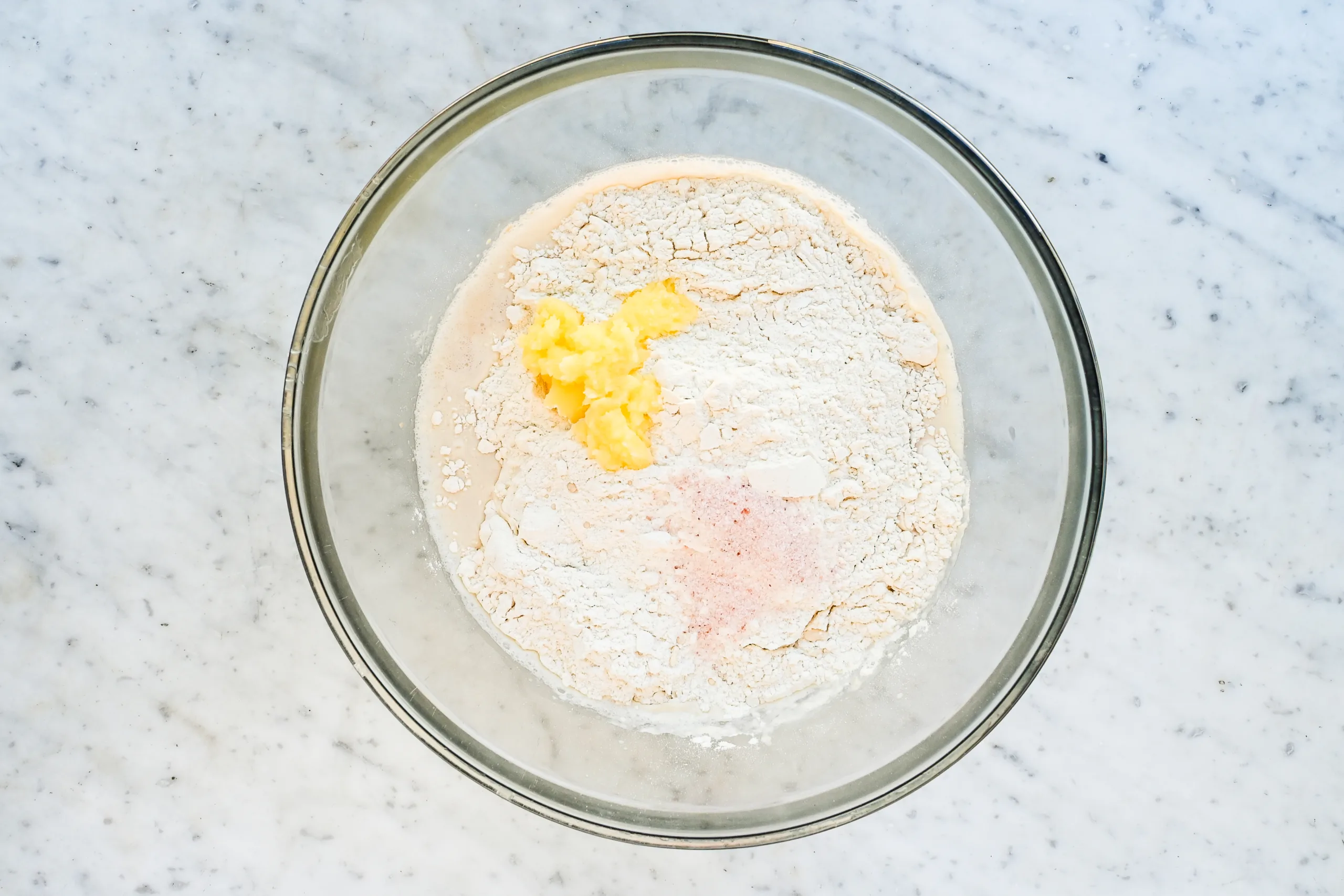Sourdough focaccia dough ingredients in a glass mixing bowl, including flour, salt, melted butter, and yeast mixture.