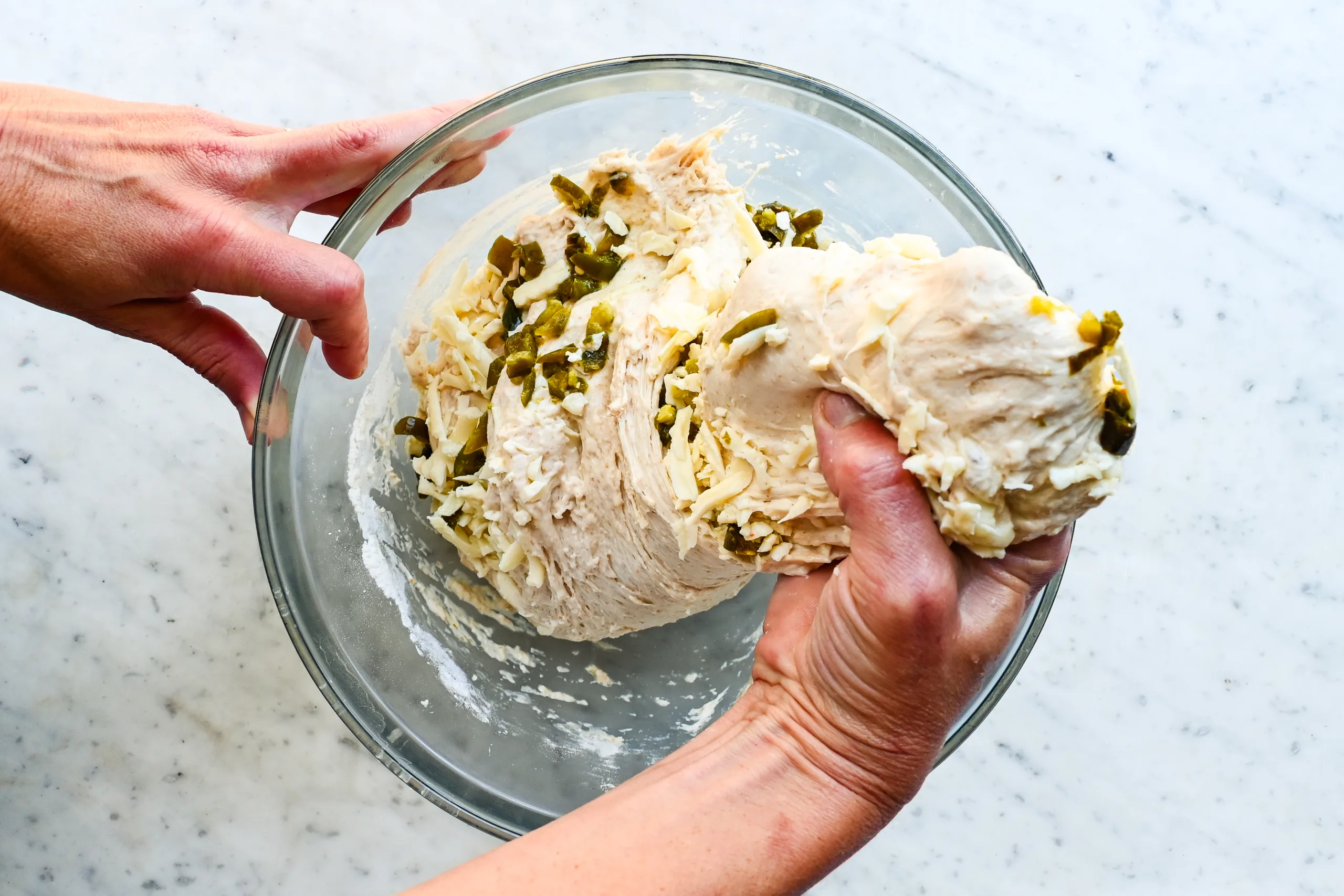 Hands stretching and folding jalapeño cheddar sourdough focaccia dough in a glass mixing bowl.