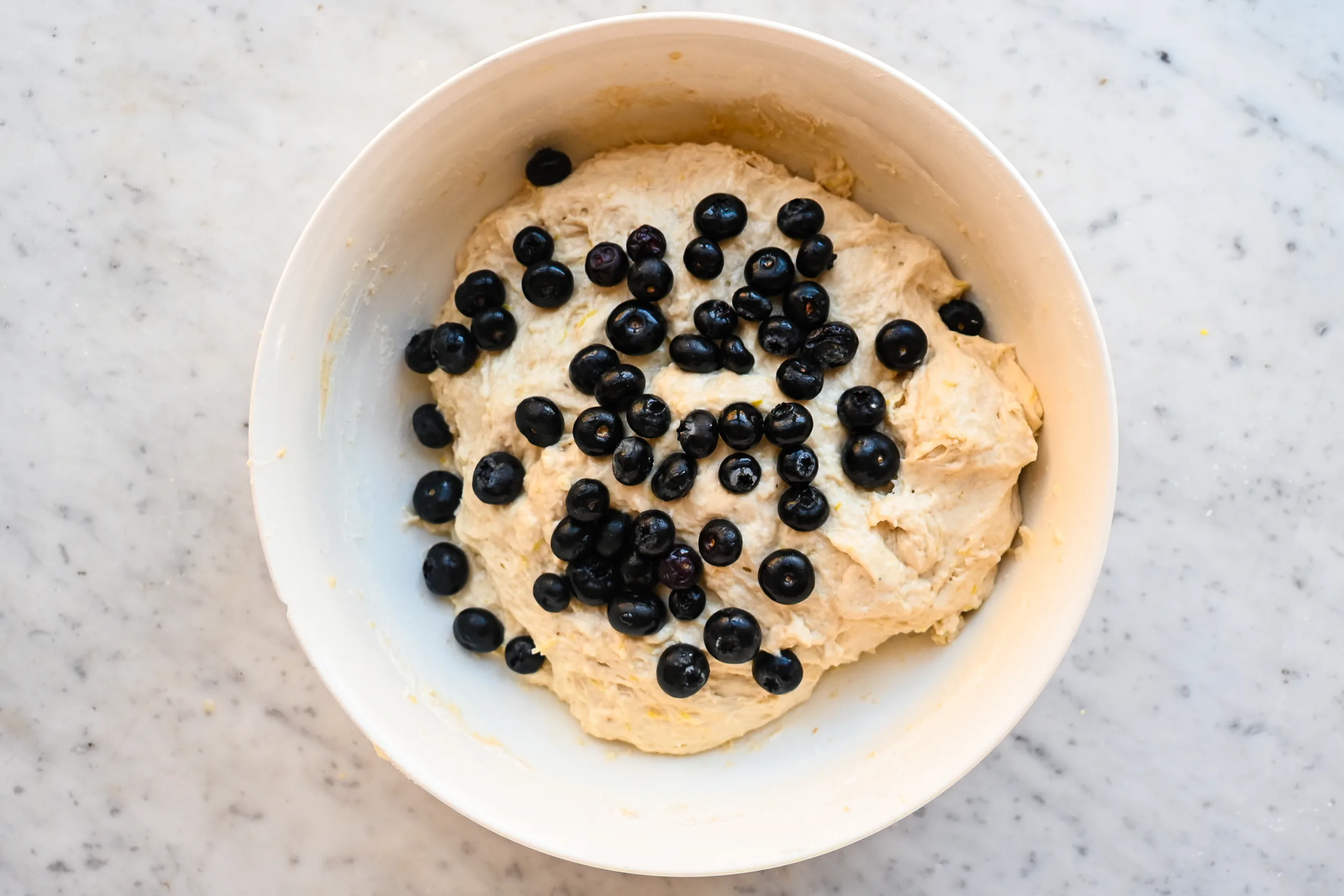 Fresh blueberries scattered over lemon sourdough focaccia dough in a white mixing bowl.