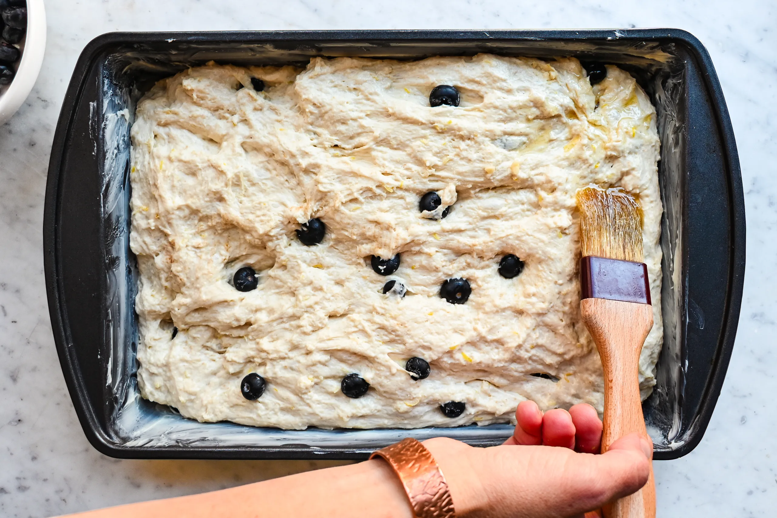 Garlic-style pastry brush brushing melted butter over lemon blueberry sourdough focaccia dough before topping.