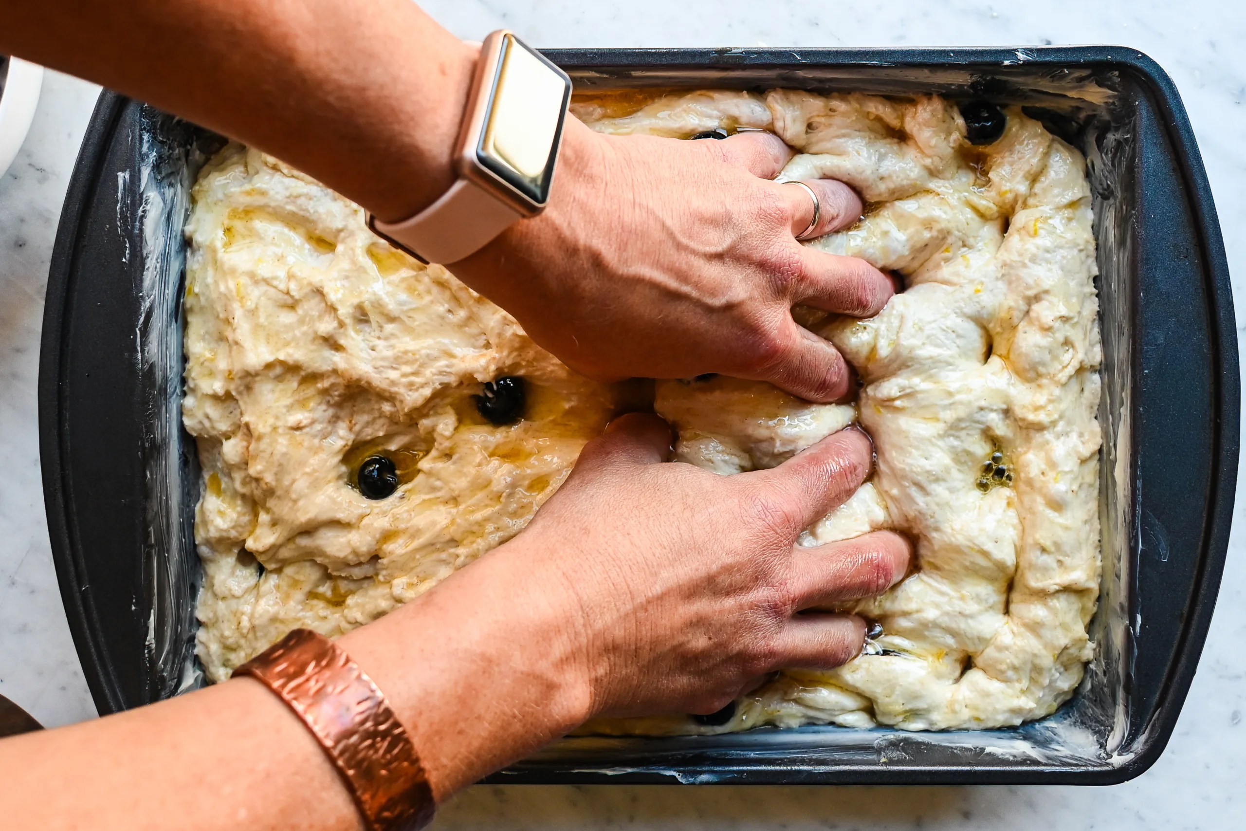 Hands dimpling lemon blueberry sourdough focaccia dough in a buttered baking pan.