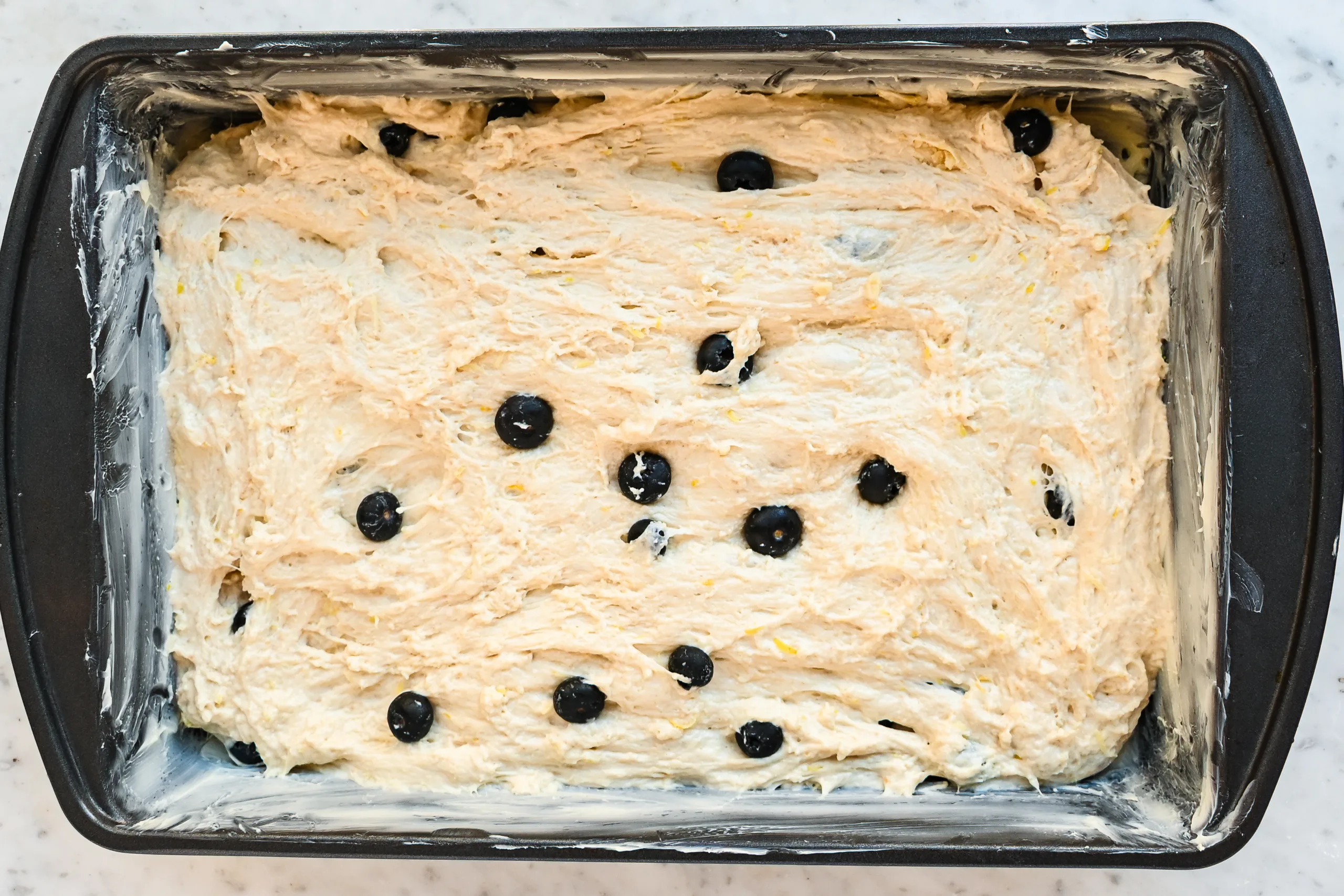 Lemon blueberry sourdough focaccia dough spread into a buttered 9x13-inch baking pan with blueberries throughout.