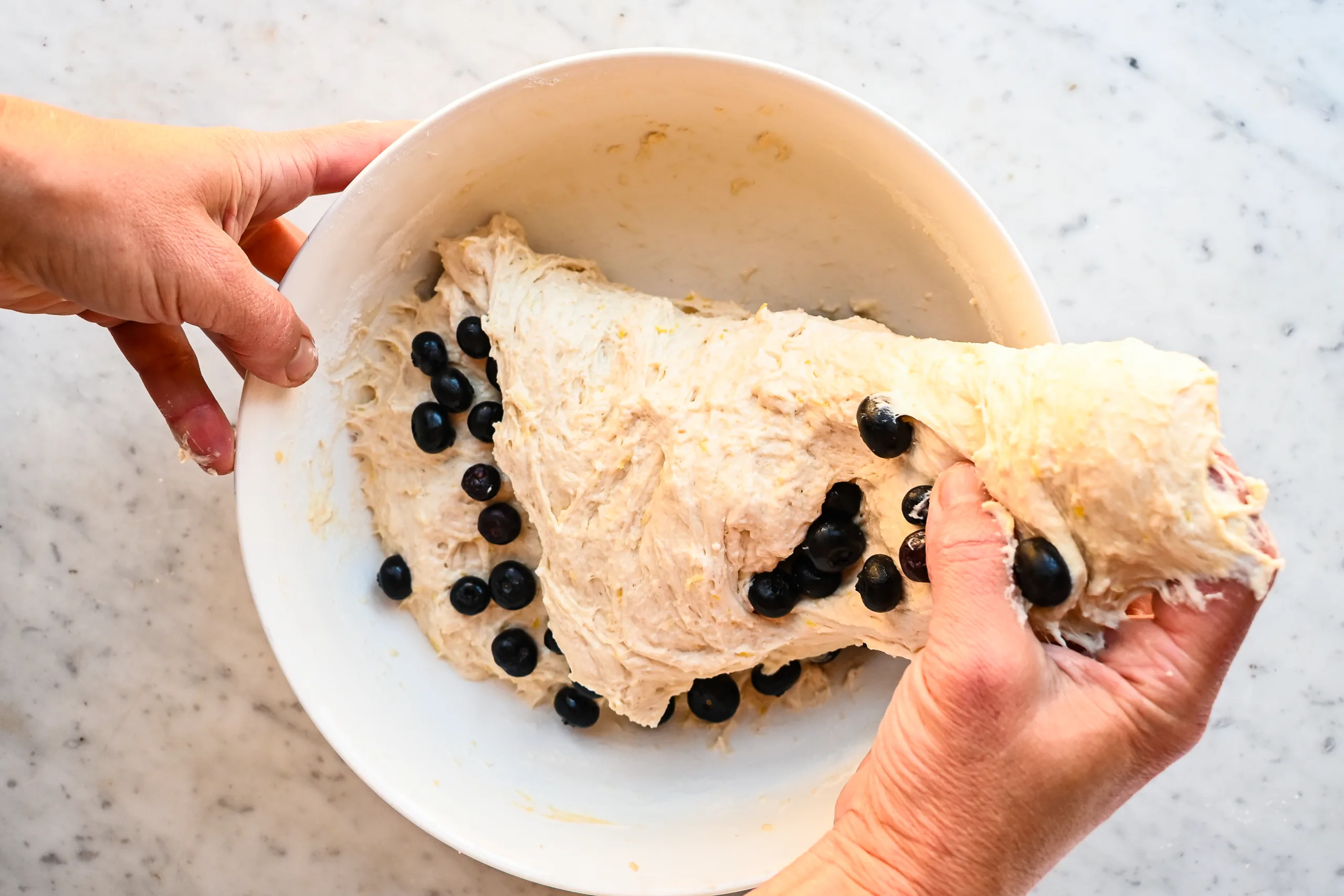 Hands stretching and folding blueberries into lemon sourdough focaccia dough.