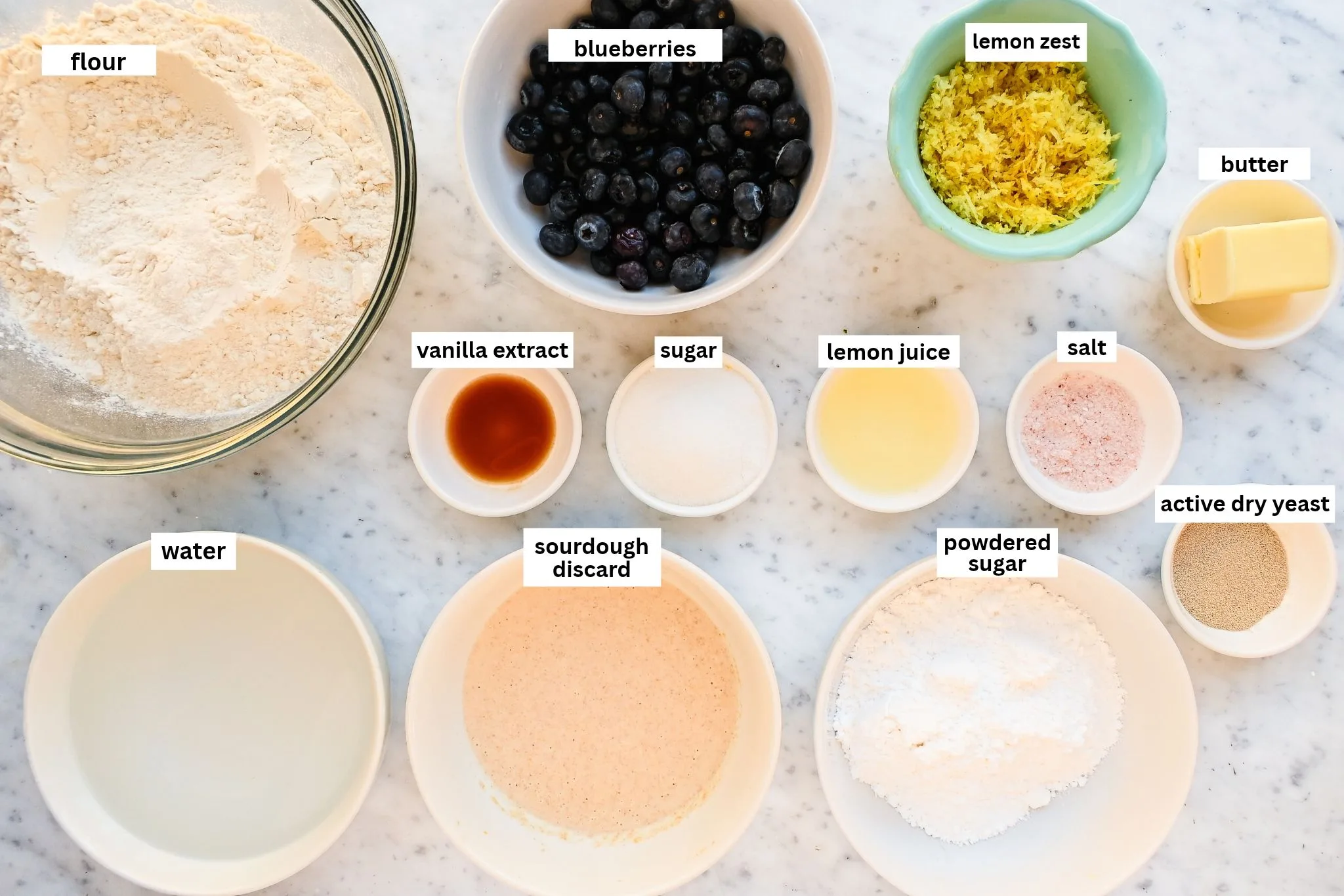 Ingredients for lemon blueberry sourdough focaccia arranged on a marble counter, including flour, blueberries, lemon zest, butter, sourdough discard, sugar, salt, vanilla, and yeast.