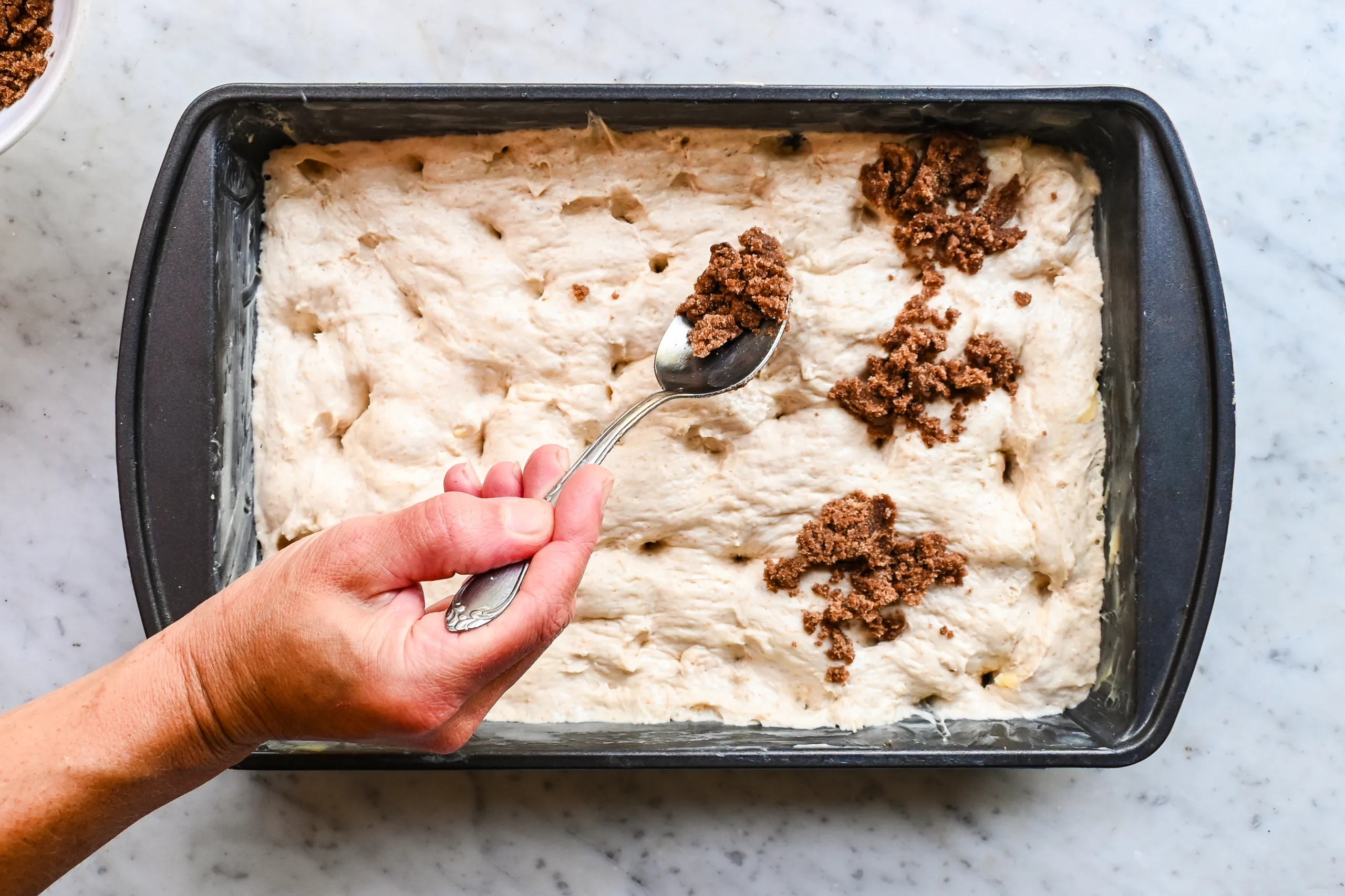 Cinnamon sugar topping being spooned over dimpled sourdough focaccia dough.