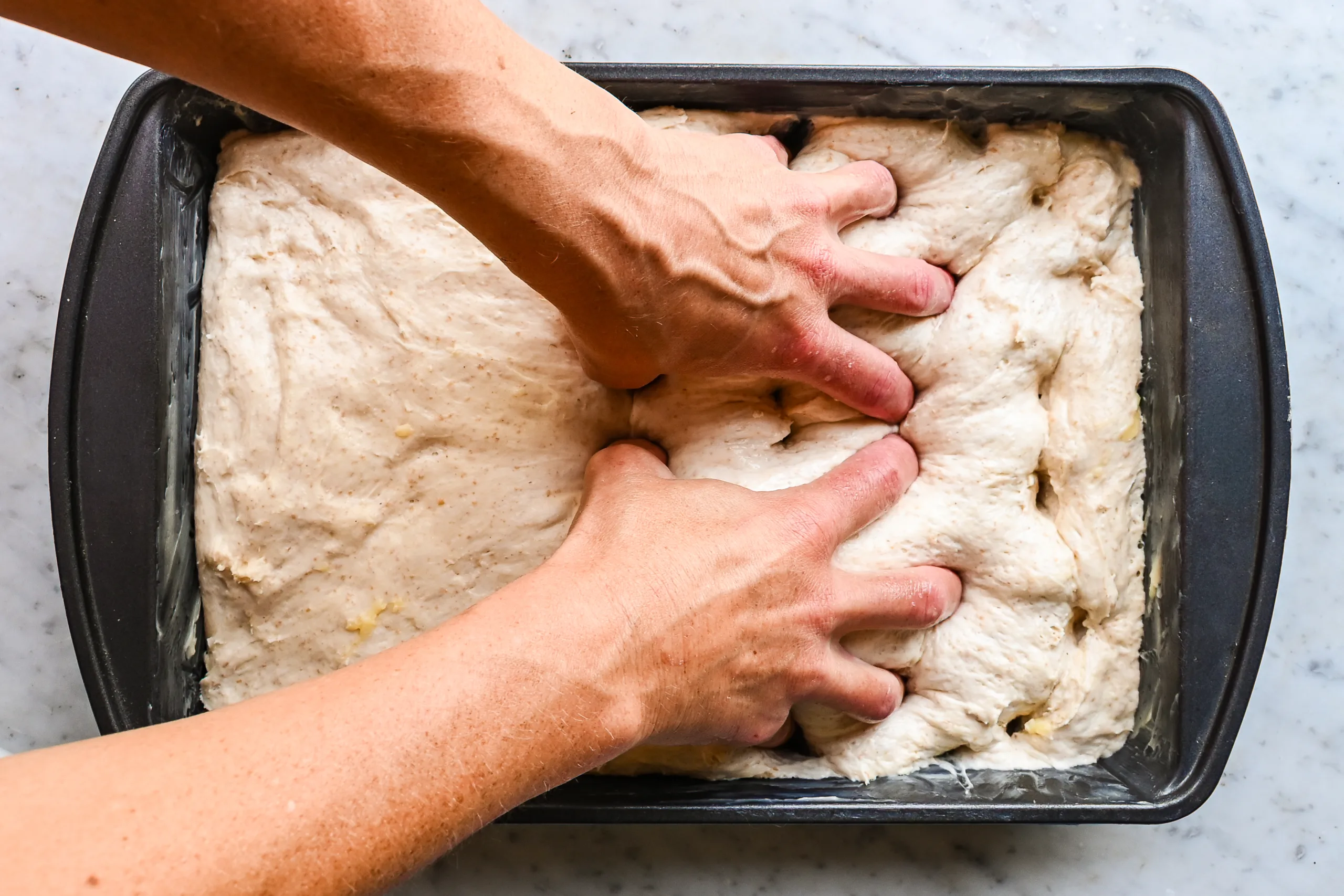Hands dimpling focaccia dough in a buttered baking pan.