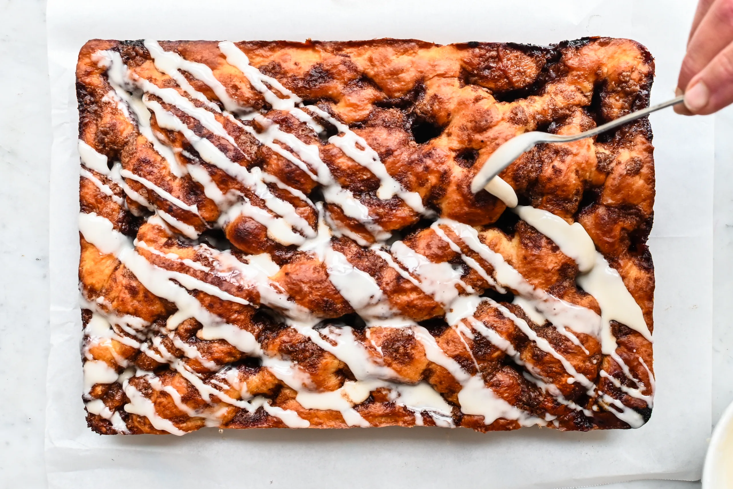 Vanilla glaze being drizzled over baked cinnamon roll focaccia.
