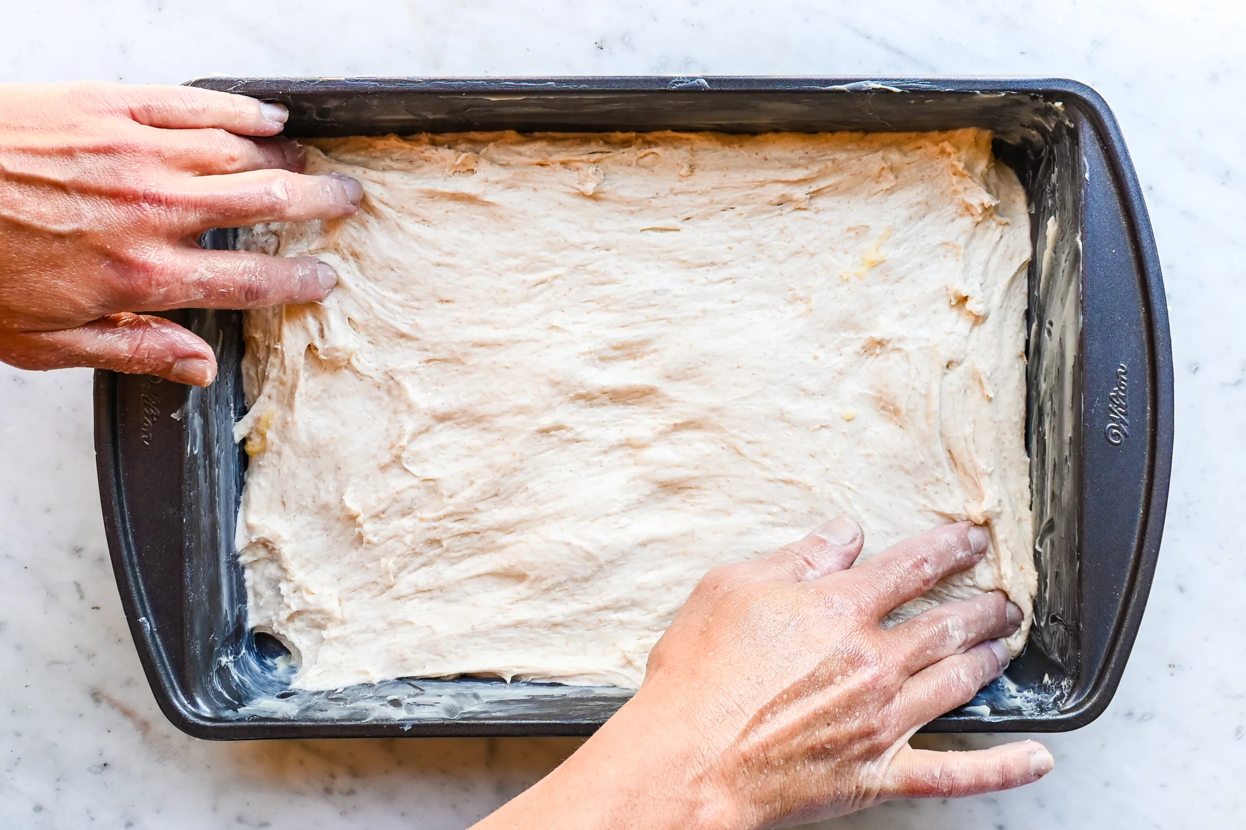 Sourdough focaccia dough being stretched into a buttered 9x13-inch baking pan.